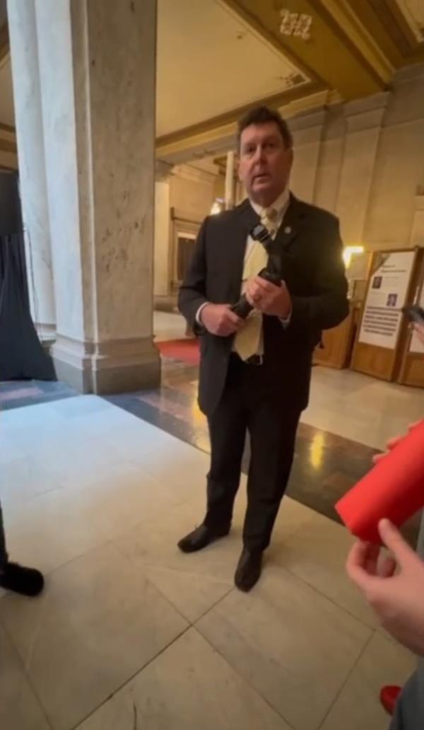 Indiana Rep. Jim Lucas holding a gun in a holster while wearing a suit and standing in front of micropho<em></em>nes at the Statehouse in Indianapolis