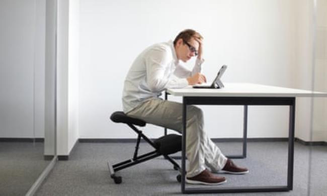 A young man is bent over his tablet in his office, seated on kneeling chair