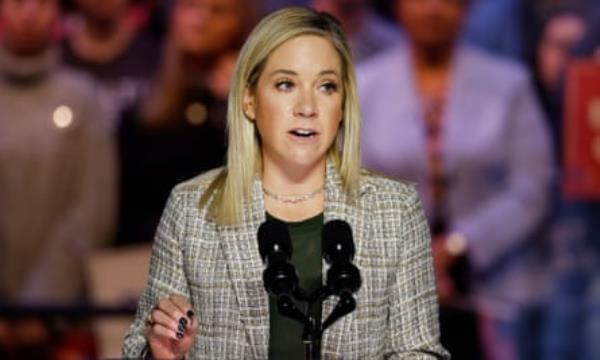 Young white woman, blond hair, gray blazer, speaks into double microphones.