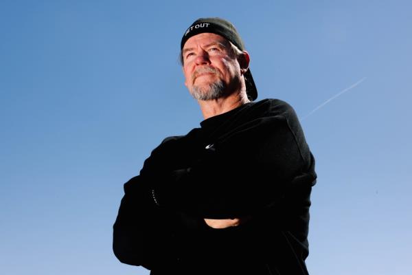 Flash Shelton wears all black, and a cap on backwards, as he stares off into blue sky