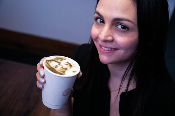 Nate Myles of Harlem, in a white button down shirt with a ring on a silver necklace around his neck, wearing a beaded bracelet and an earring, snapping a selfie at Moka Matcha Cafe, with colorful are of different coffee cups on the wall behind him