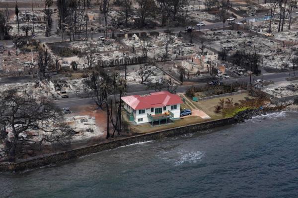 A two-story house, sporting white walls and a red roof, stands tall amid a devastated Lahaina.  