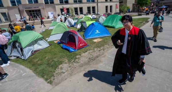 A student in a graduation cap and gown walks by the protest encampment Monday, May 6, 2024 at the University of Wisconsin