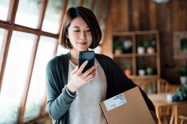 Smiling young Asian woman shopping o<em></em>nline with smartphone on hand, receiving a delivered parcel by home delivery service.