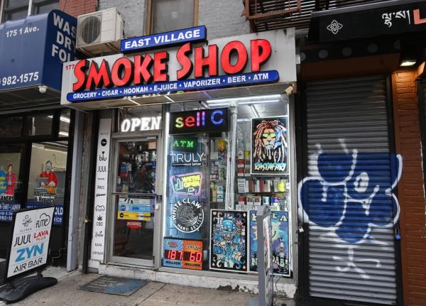 A shop branded East Village Smoke Shop. It has advertising in the window for Rastafari with a logo of a lion with dreads, White Claw hards seltzer, and has a lit-up signing which says 