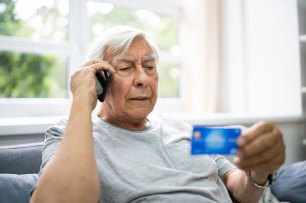 A man holding a credit card and a phone to his ear, representing elder scam call and senior pension finance fraud