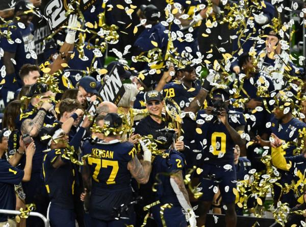 Michigan Wolverines head coach Jim Harbaugh holding Natio<em></em>nal Champio<em></em>nship trophy with celebration in the background.