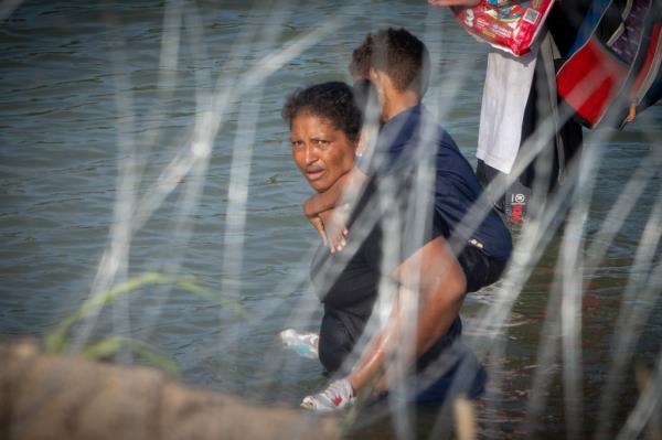 A woman holds a child on her back as migrants walk near co<em></em>ncertina wire in the water along the Rio Grande border