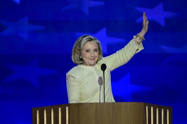 Hillary Rodham Clinton delivering remarks at the 2024 Democratic Natio<em></em>nal Co<em></em>nvention in Chicago, standing at a podium with her hand raised