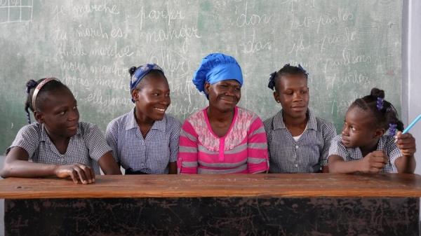 Haitian woman in pink shirt and blue headdress sits with four girls in school uniforms against a blackboard